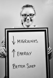 A woman is smiling behind a sign with the word migraines and energy written on it.