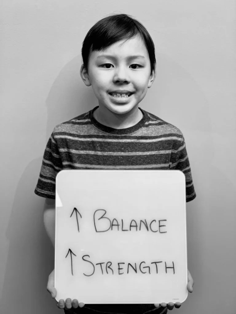 patient story - A young boy holding up a sign that says improved balance and strength- neuro-structural chiropractic adjustment results for pediatrics