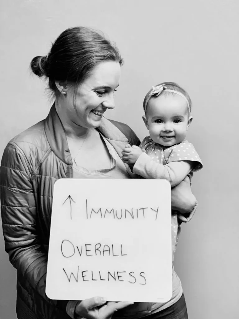patient story - A woman holding a baby and a sign that says improved immunity and overall wellness- neuro-structural chiropractic adjustment results for pediatrics