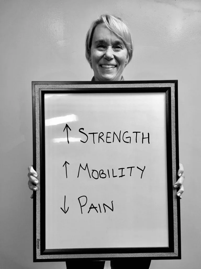 patient story - A woman holding up a sign that says improved strength, improved mobility, and reduced pain - neuro-structural chiropractic adjustment results for adults