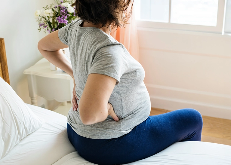woman sitting on the bed with with a big belly and hands supporting her back- chiropractic care for pregnancy in the third trimester
